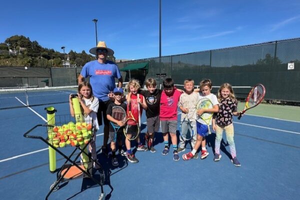 A group of children and an adult stand on a tennis court with rackets, next to a basket of tennis balls, under a clear blue sky.