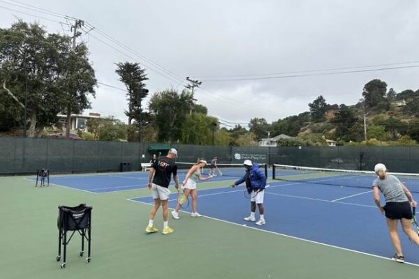 Four people stand on an outdoor tennis court, with two engaged near the net and two others walking nearby; tennis equipment is visible on the court.