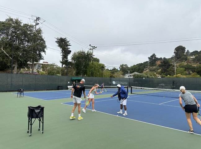 Four people stand on an outdoor tennis court, with two engaged near the net and two others walking nearby; tennis equipment is visible on the court.