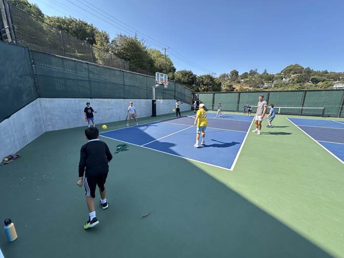 A group of children and adults play a ball game on a tennis court under a clear sky, with some watching and others actively participating.