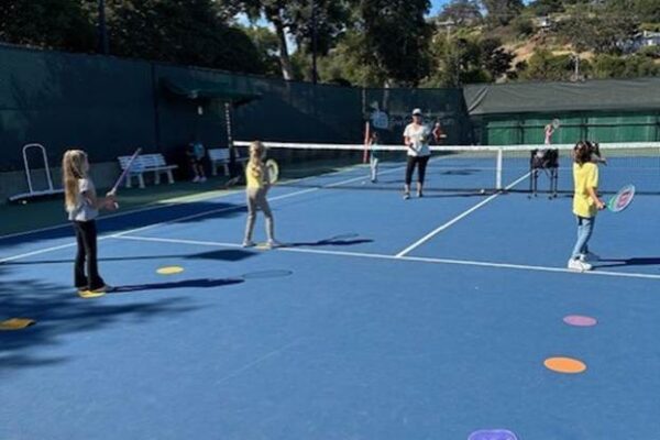 Four children stand on a blue tennis court holding rackets, with an adult instructing them; colored spots mark positions on the court.