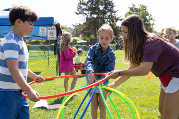 Children and an adult assemble a structure with colorful plastic hoops on a grassy field, with other children and outdoor equipment in the background.