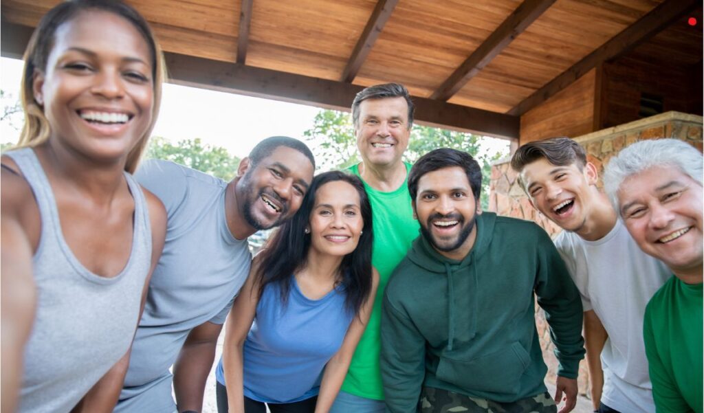A diverse group of seven adults stands close together under a wooden roof, smiling at the camera in a casual, outdoor setting.