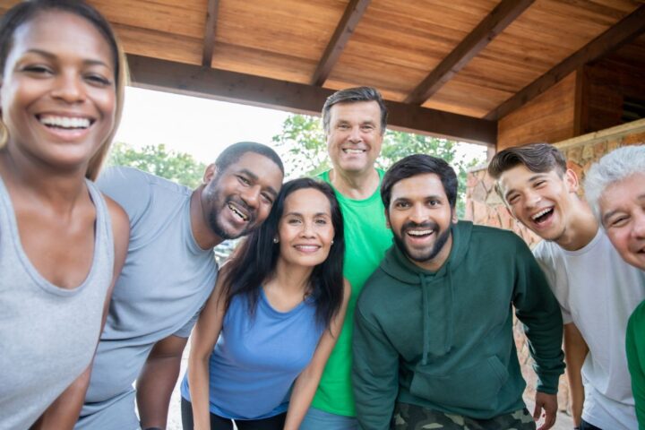 A diverse group of seven adults stands close together under a wooden roof, smiling at the camera in a casual, outdoor setting.