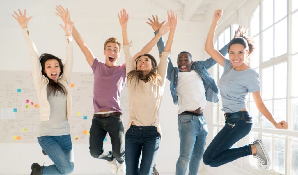 Five people stand in a bright room, jumping with arms raised and smiling, with large windows and a whiteboard in the background.