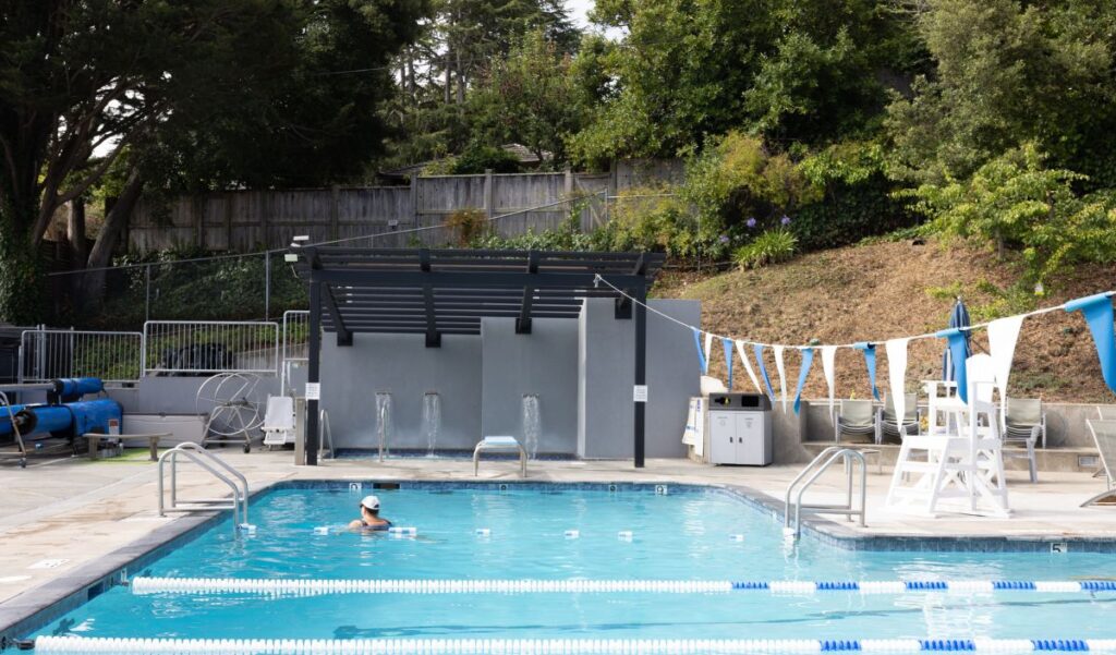 A person swims alone in an outdoor pool with lane dividers, a lifeguard chair, and trees and shrubs in the background.