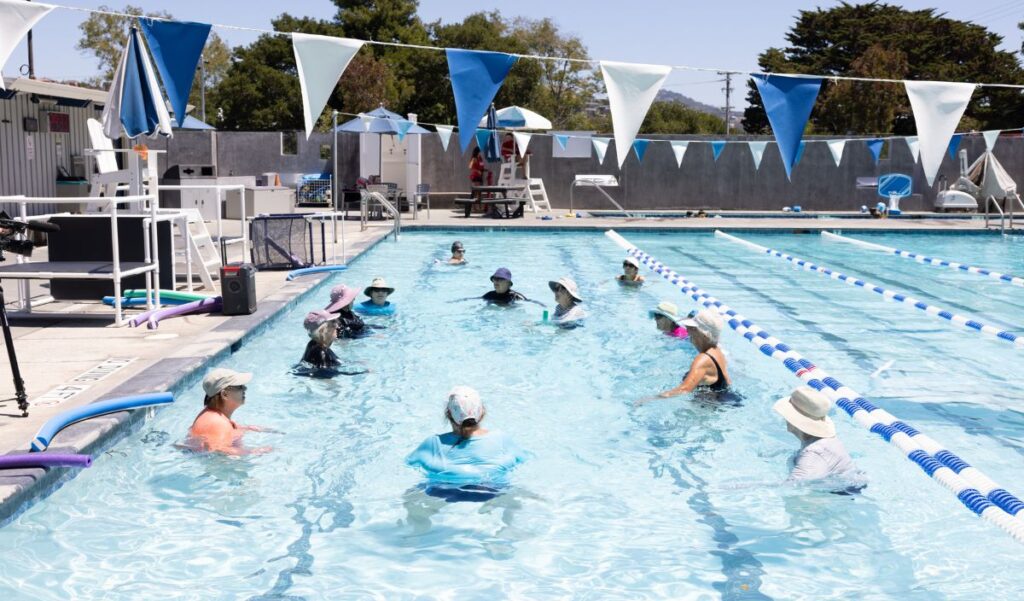 A group of people wearing hats and sun-protective clothing participate in a water exercise class in an outdoor swimming pool.