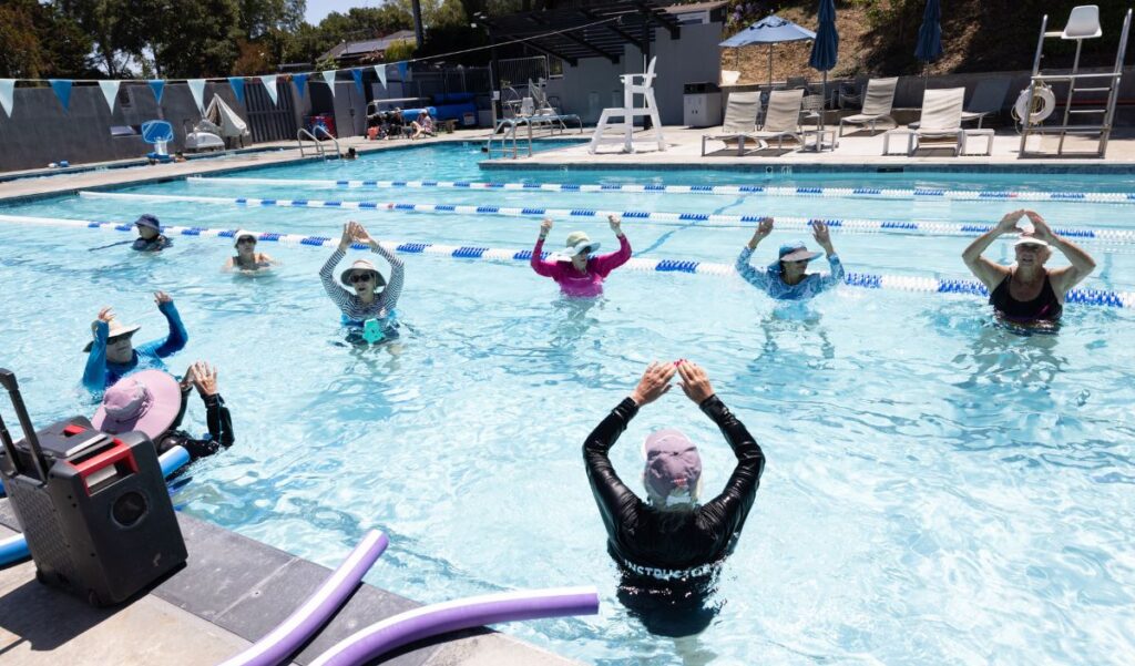A group of people participate in a water aerobics class in an outdoor swimming pool, following an instructor at the front.