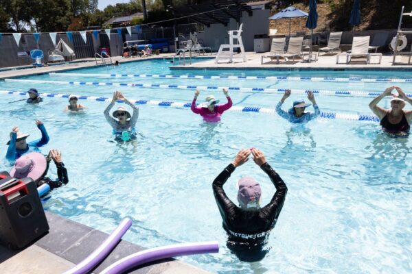 A group of people participate in a water aerobics class in an outdoor swimming pool, following an instructor at the front.