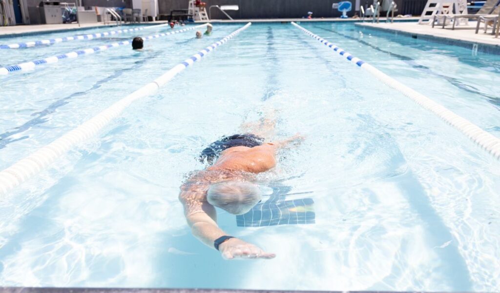 Person swimming freestyle in an outdoor lane pool, wearing a swim cap and goggles; other swimmers are visible in adjacent lanes.