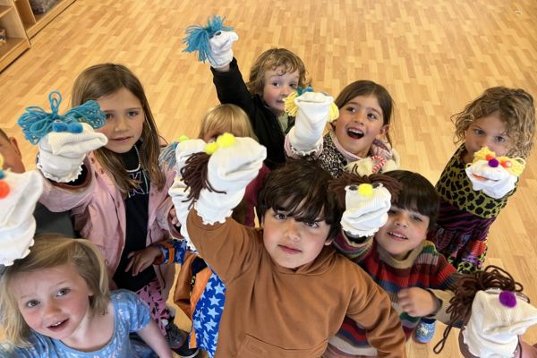 A group of young children stand in a circle holding up handmade sock puppets, smiling and looking up at the camera in a brightly lit room with a wooden floor.
