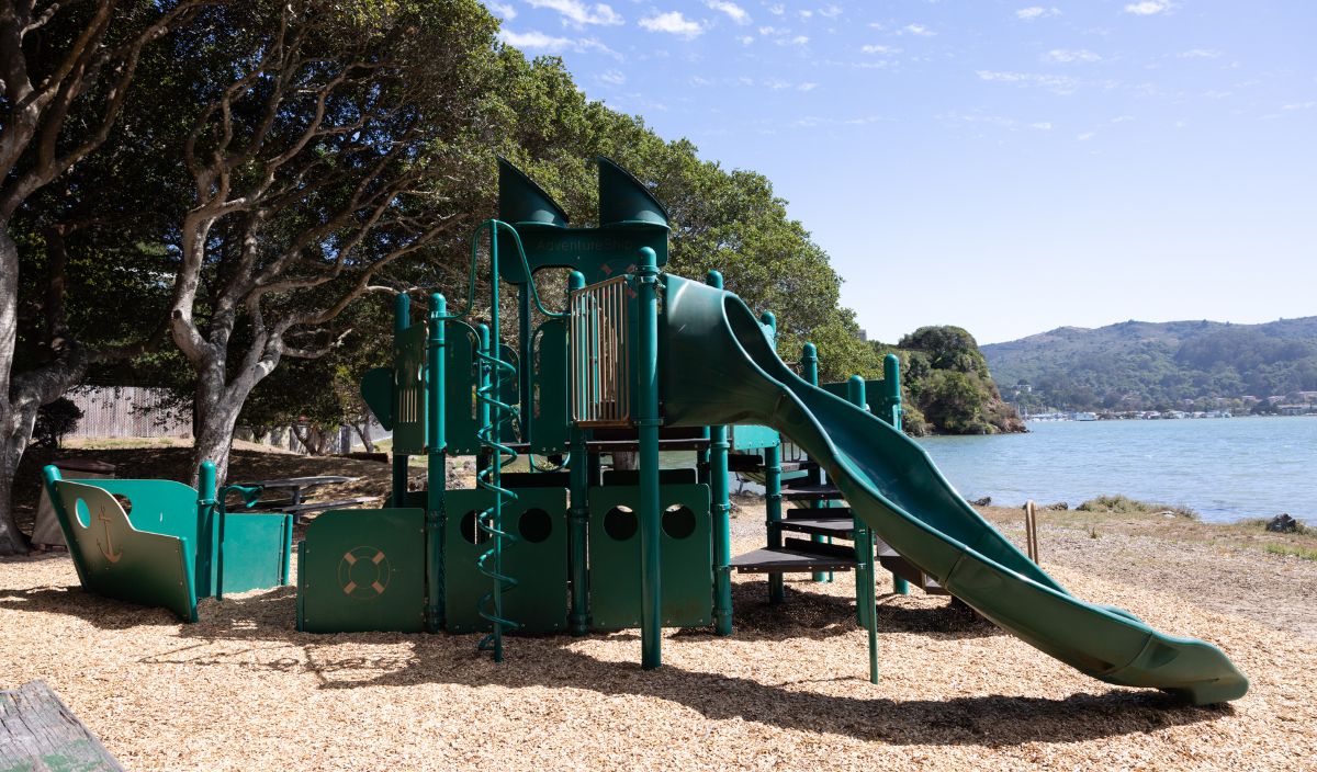 Green playground structure resembling a ship with slides and climbing features, situated near trees and a body of water under a blue sky.