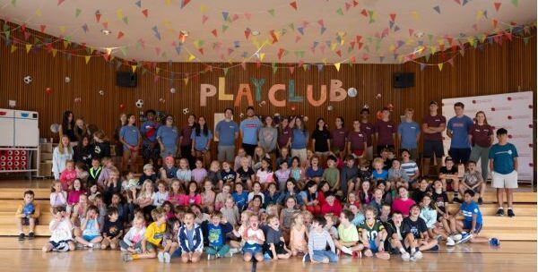 Large group of children and adults posed together in a gymnasium decorated with colorful banners and a “PLAY CLUB” sign on the back wall.
