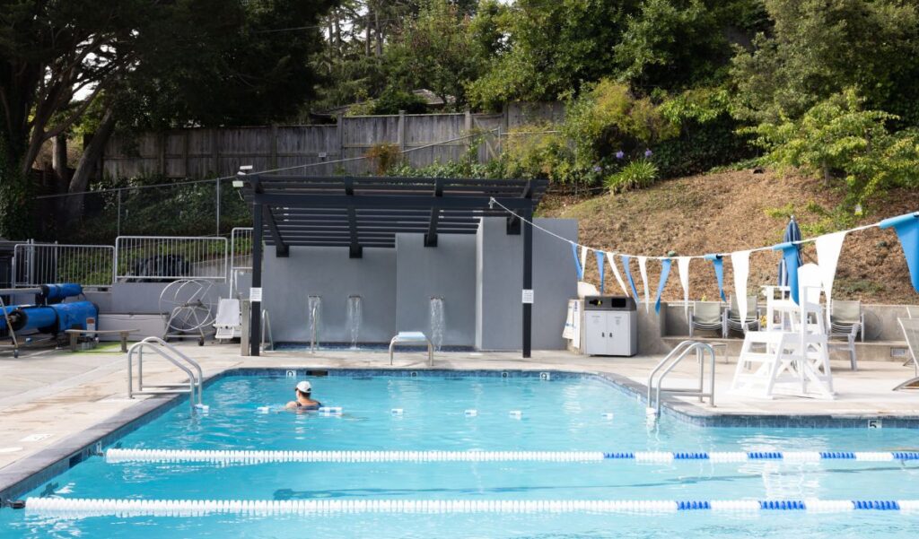 A single swimmer is in a lap lane of an outdoor pool, which is surrounded by trees, equipment, and a shaded structure. Blue and white flags hang above the water.