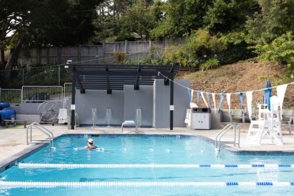 A single swimmer is in a lap lane of an outdoor pool, which is surrounded by trees, equipment, and a shaded structure. Blue and white flags hang above the water.
