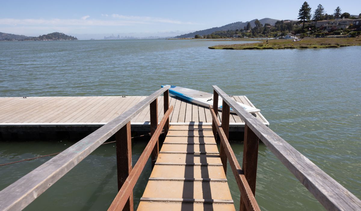 A wooden walkway leads to a dock over calm water, with a paddleboard resting on the edge and houses visible on the distant shoreline.