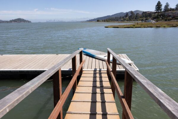Wooden ramp leading to a floating dock on a calm body of water, with a paddleboard nearby and hills in the background under a clear sky.