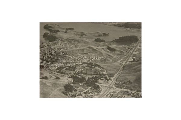 Aerial view of a suburban area with houses, open land, a roadway, and a body of water in the background.