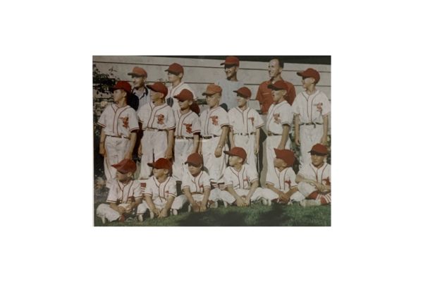 A youth baseball team in white uniforms and red caps poses for a group photo outdoors with their coach.