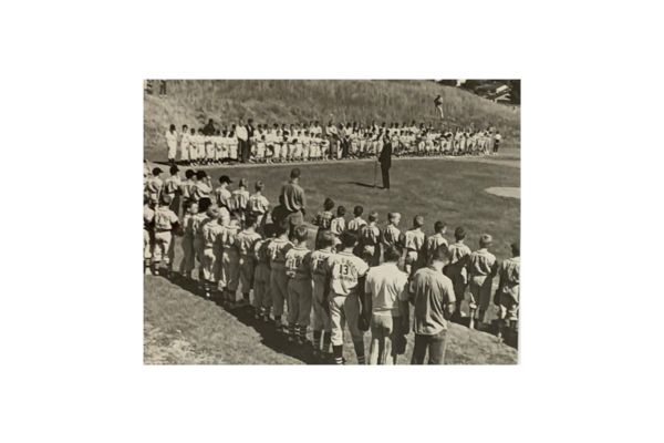 A group of baseball players stand in uniform lines on a field, facing a person speaking in front of another group, with grassy hills in the background.