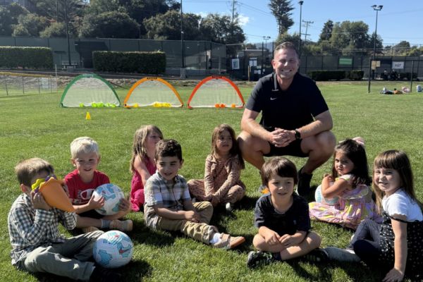 A group of young children and an adult coach are sitting on grass with soccer balls in front of small soccer goals on a sunny day.