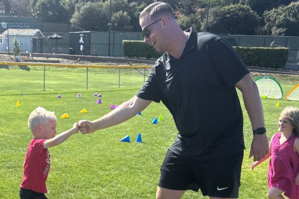 An adult wearing sunglasses and a black outfit gives a fist bump to a young child on a grassy field with colored cones and two other children nearby.