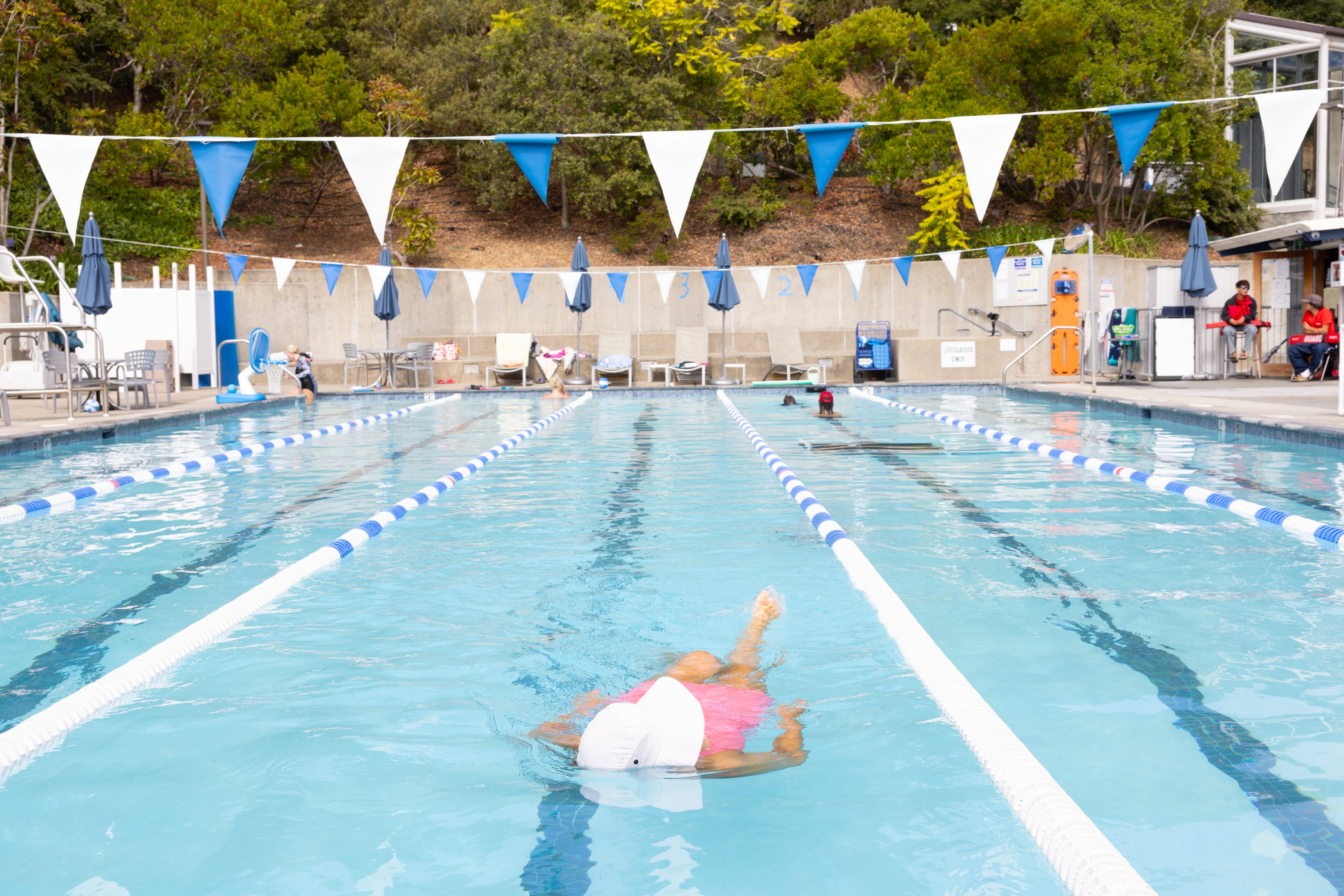 Person swimming freestyle in an outdoor lap pool with blue and white lane dividers; other people sit or stand around the poolside. Trees and umbrellas are visible in the background.