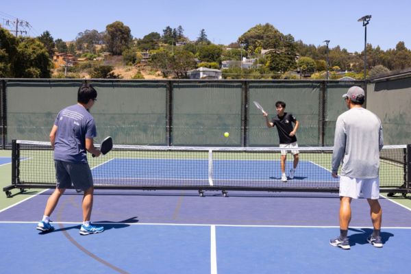 Three men are playing doubles pickleball on an outdoor court, with one player preparing to hit the ball over the net.
