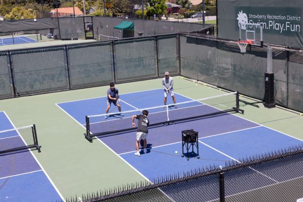 Four people play pickleball on an outdoor court, with two on each side of the net. A ball cart is on the court, and a large sign reads "Strawberry Recreation District.