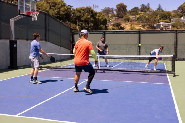 Four men play a game of pickleball on an outdoor court, with two players on each side of the net on a sunny day.