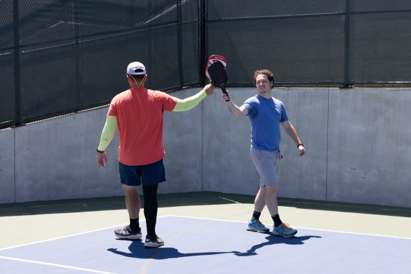 Two men on an outdoor court, one in blue and one in orange, tap their pickleball paddles together in a gesture of sportsmanship.