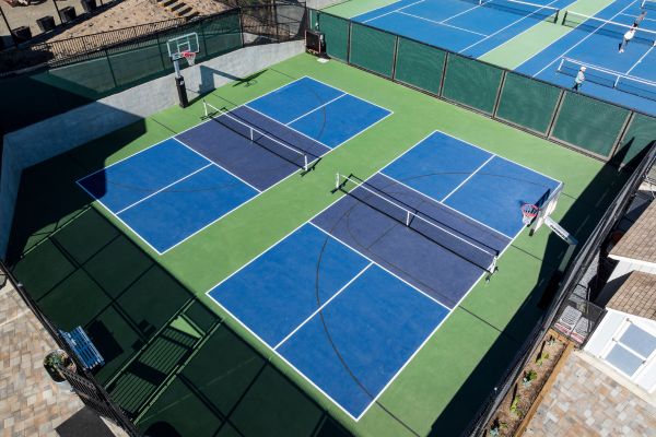 Aerial view of two pickleball courts and two basketball hoops next to a set of blue tennis courts, all surrounded by fences.
