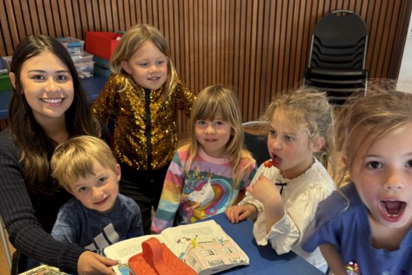 A woman and five young children sit around a table with an open book, smiling and looking at the camera in a classroom setting.