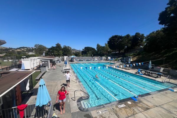 Outdoor swimming pool with lane dividers, a few people swimming, lifeguards on deck, umbrellas, and trees in the background on a sunny day.