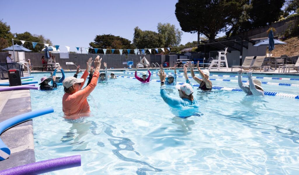 A group of people in hats and swim shirts participate in a water exercise class in an outdoor pool, raising their arms above their heads.