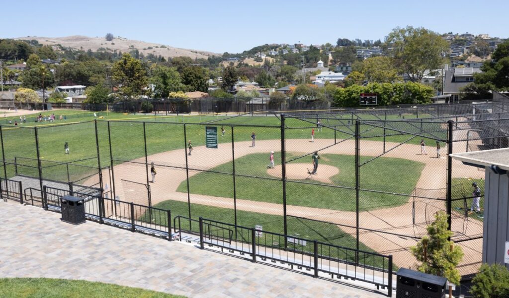 A youth baseball game is being played on a fenced field, with players on the diamond and spectators nearby; houses and hills are visible in the background.
