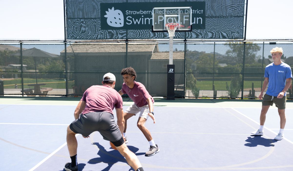 Three young men play basketball outdoors on a sunny day, with one dribbling while another defends. A sign in the background reads "Swim. Play. Fun.