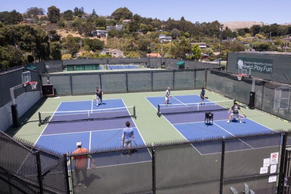 Six people are playing doubles pickleball on two adjacent outdoor courts surrounded by fencing, with trees and houses in the background.
