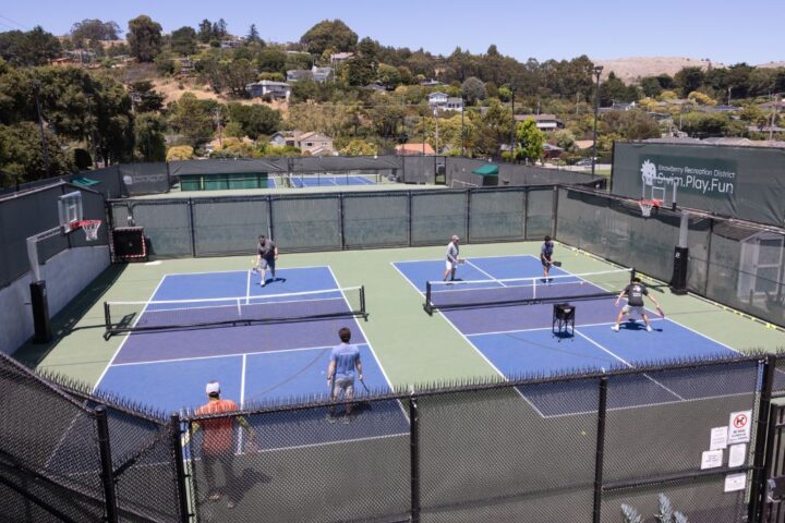 Six people are playing doubles pickleball on two adjacent outdoor courts surrounded by fencing, with trees and houses in the background.