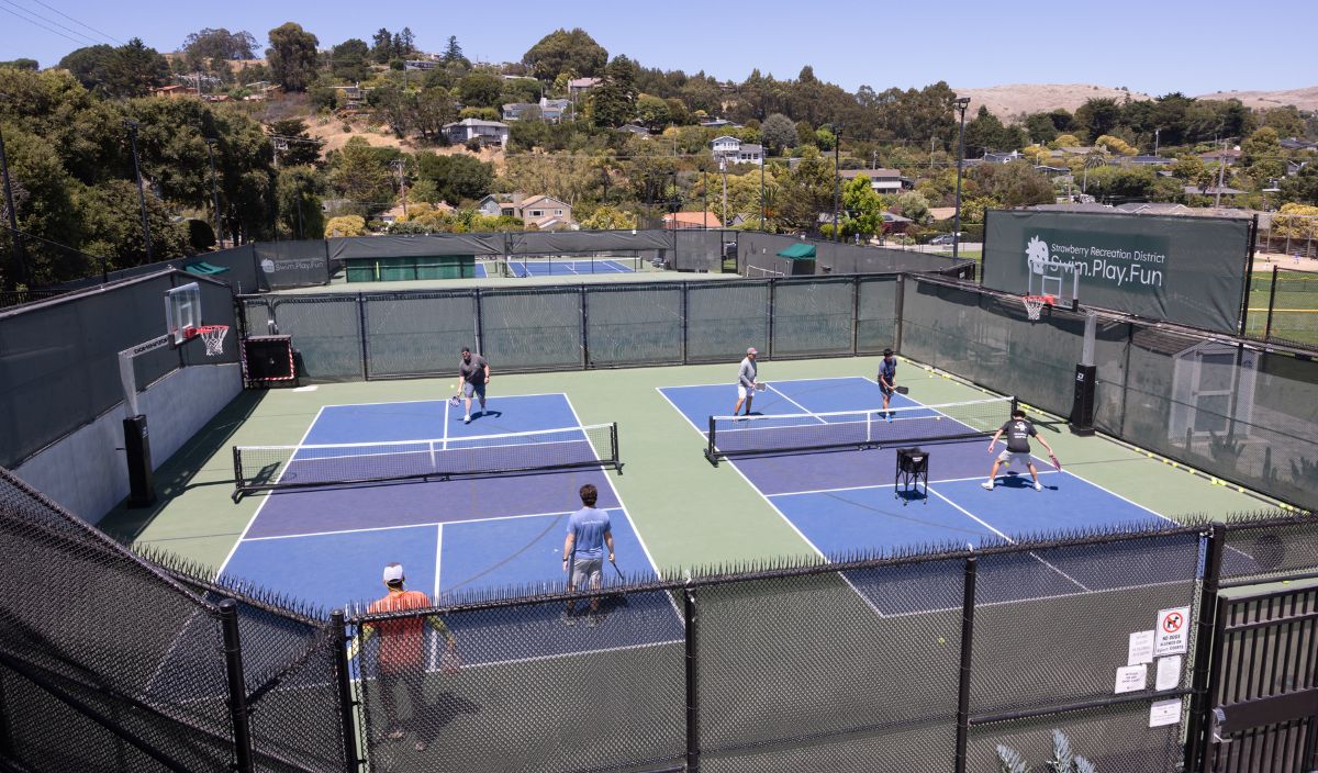 Six people are playing doubles pickleball on two adjacent outdoor courts surrounded by fencing, with trees and houses in the background.