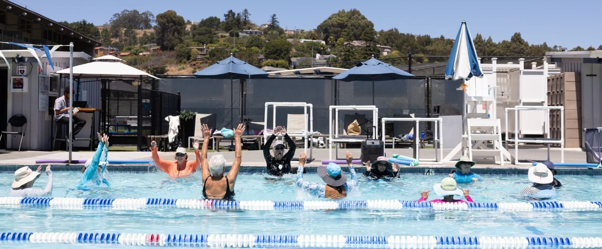 A group of people in hats participate in a water exercise class in an outdoor pool on a sunny day, with trees and buildings in the background.