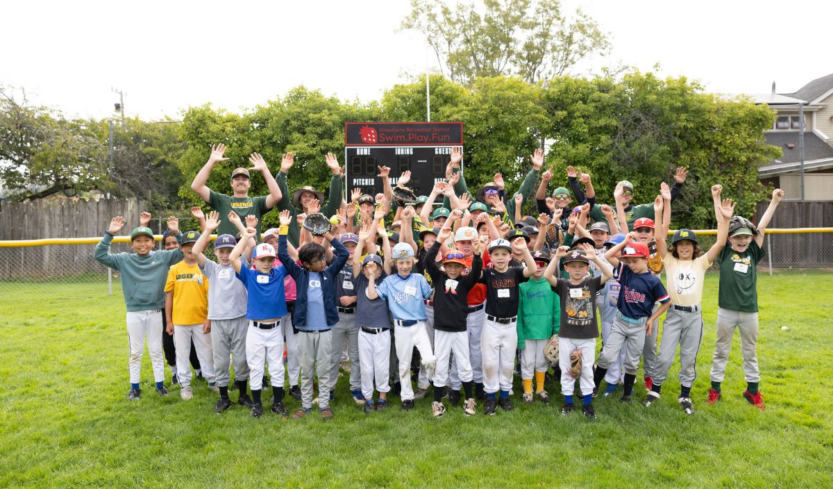 A large group of children and adults in baseball uniforms pose on a grassy field with their arms raised, standing in front of a scoreboard and trees.