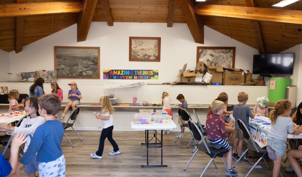 Children participate in various arts and crafts activities in a classroom with a wooden ceiling and a sign reading "AMAZING THINGS HAPPEN HERE" on the wall.