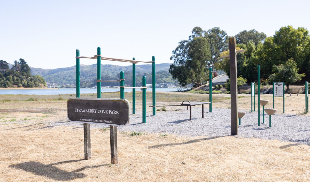 Outdoor fitness equipment and a sign reading "Strawberry Cove Park" are set on a dry grass area near a body of water, with trees and hills in the background.