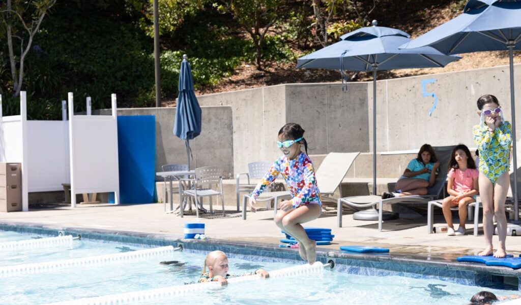 A child wearing goggles jumps into a swimming pool while another child and two adults watch from poolside lounge chairs under umbrellas.