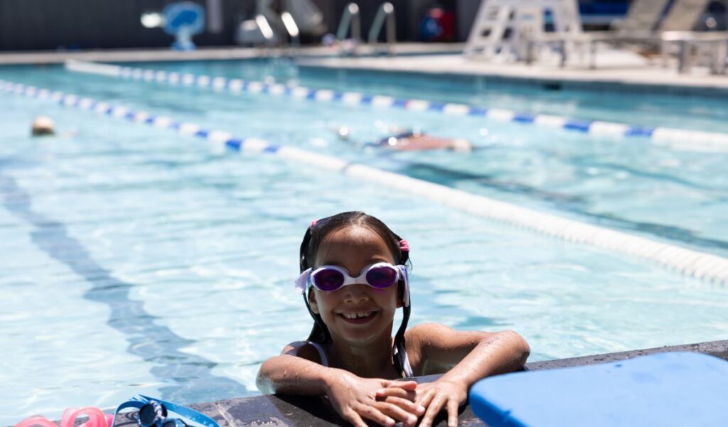 A young girl wearing swim goggles smiles while holding onto the edge of an outdoor swimming pool, with lane dividers and other swimmers in the background.