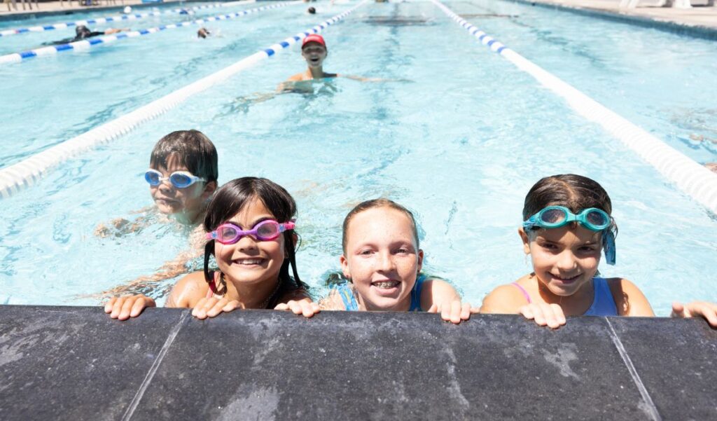 Four children in goggles smile at the edge of a swimming pool, with another child swimming in the lane behind them.