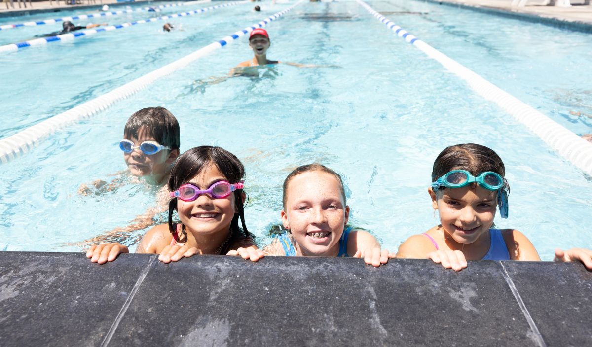 Four children in goggles smile at the edge of a swimming pool, with another child swimming in the lane behind them.