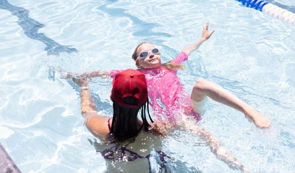 A swim instructor in a red cap assists a child wearing pink swimwear and goggles as she floats on her back in a swimming pool.
