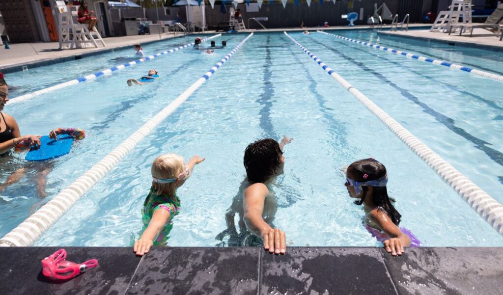 An adult teaches two children how to swim in a pool, with the children holding onto the pool edge and other people swimming in designated lanes.